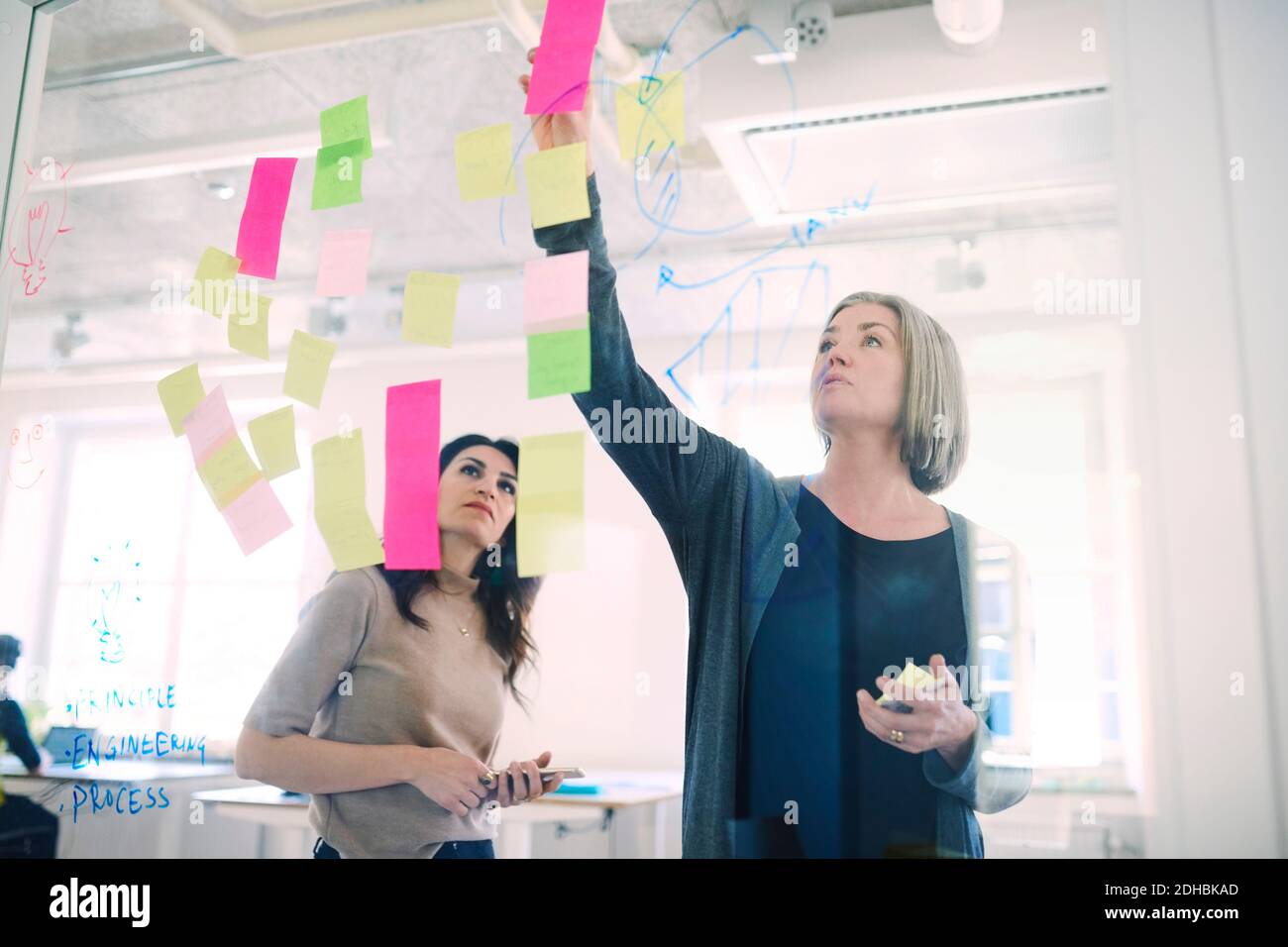 Ingegnere femminile che discute con la collega sulle note adesive sul vetro in ufficio creativo Foto Stock