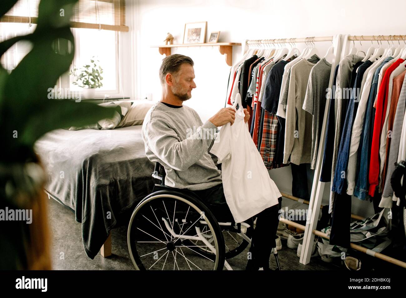Vista laterale di un uomo maturo per disabili che tiene una t-shirt bianca mentre seduta su sedia a rotelle presso il portabiti a casa Foto Stock