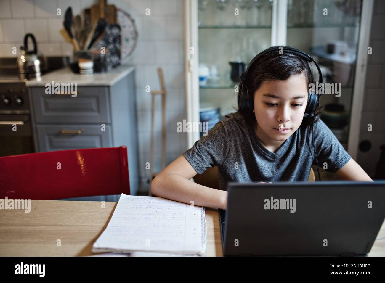 Ragazzo che indossa le cuffie mentre usa il computer portatile durante i compiti a casa Foto Stock