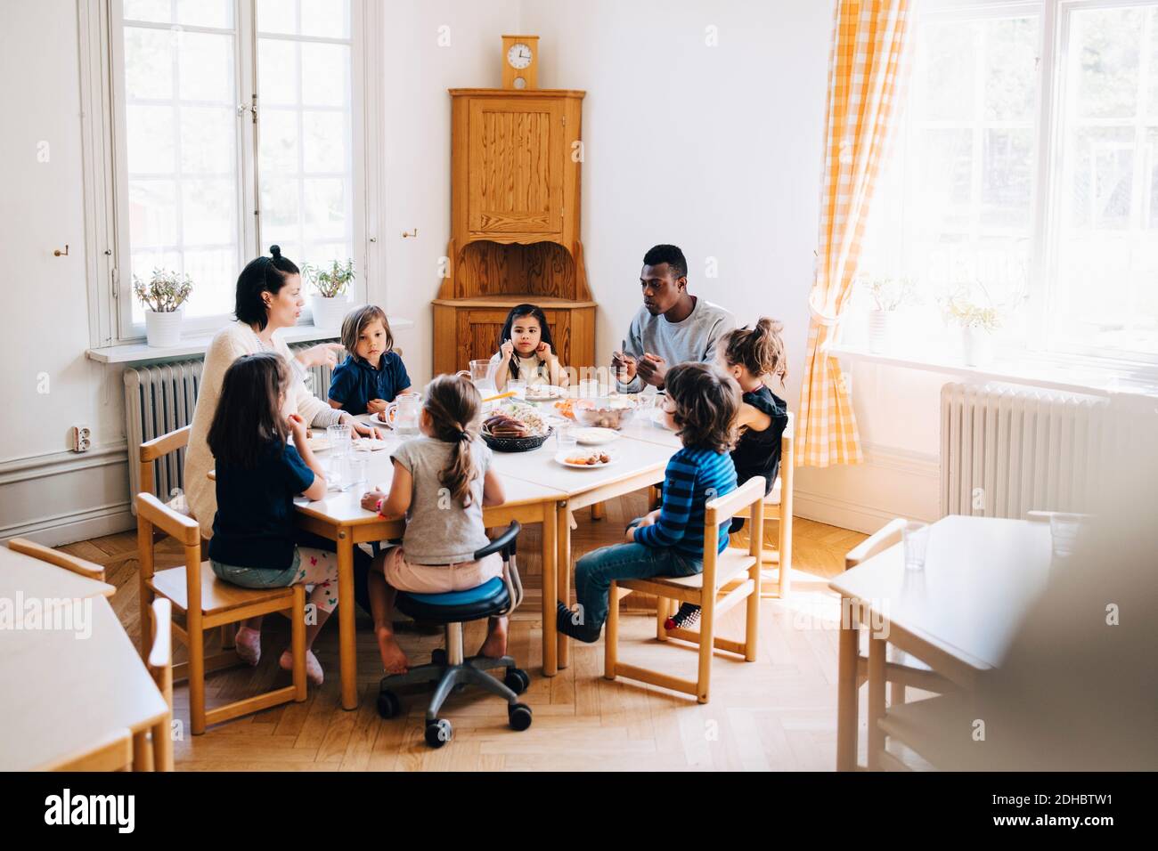 Insegnanti e studenti che mangiano il pranzo al tavolo in una scuola materna Foto Stock