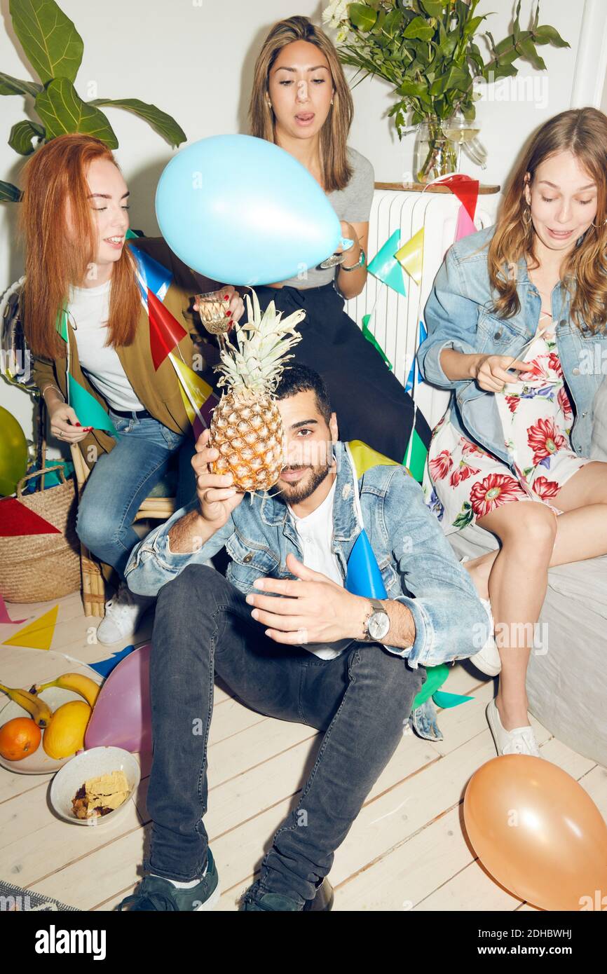 Ritratto di giovane uomo che tiene l'ananas mentre si siede contro la femmina amici che si divertano con la festa a casa Foto Stock