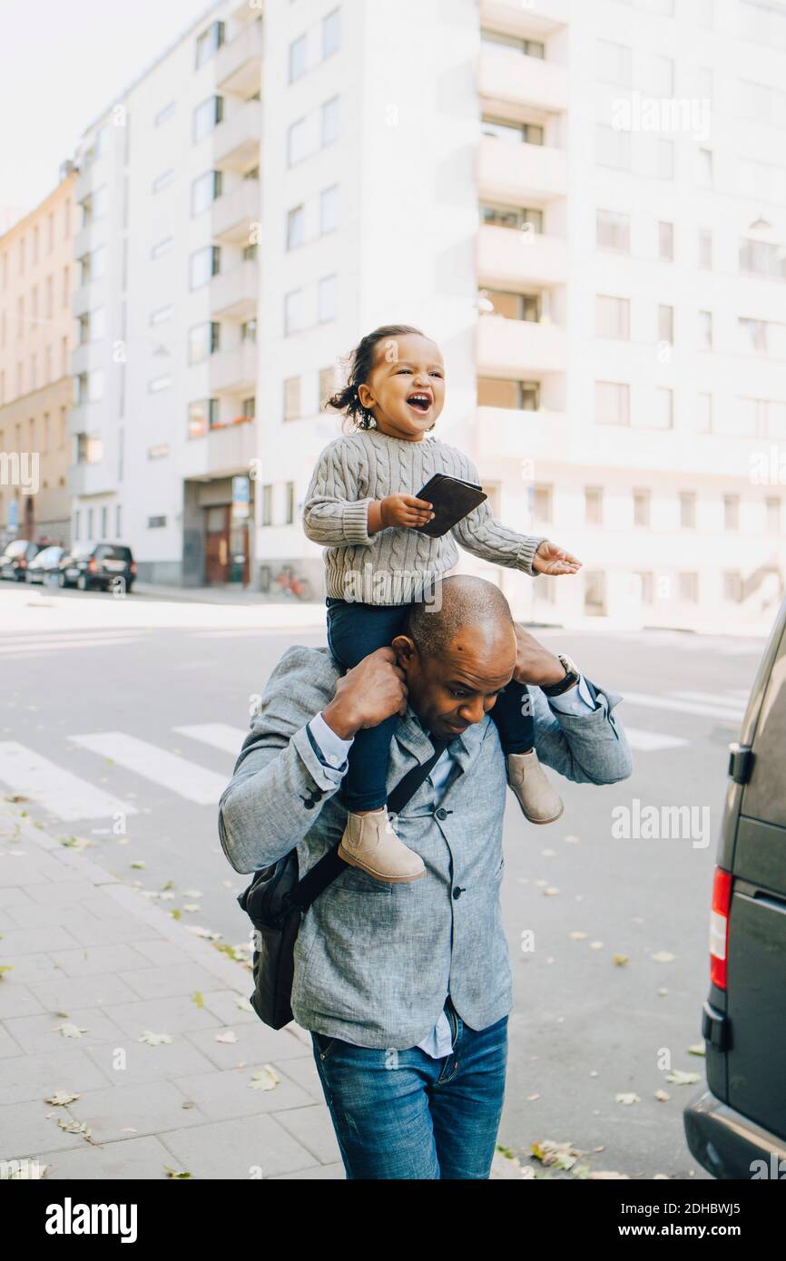 Padre che porta con sé una figlia allegra che tiene lo smartphone mentre si cammina sentiero in città Foto Stock