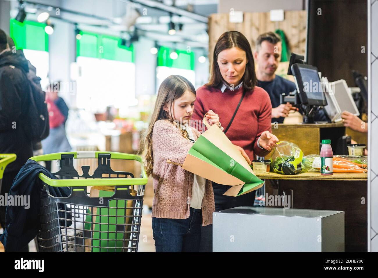 Figlia che tiene il sacchetto di carta mentre si è levati in piedi dalla madre al checkout banco nel supermercato Foto Stock