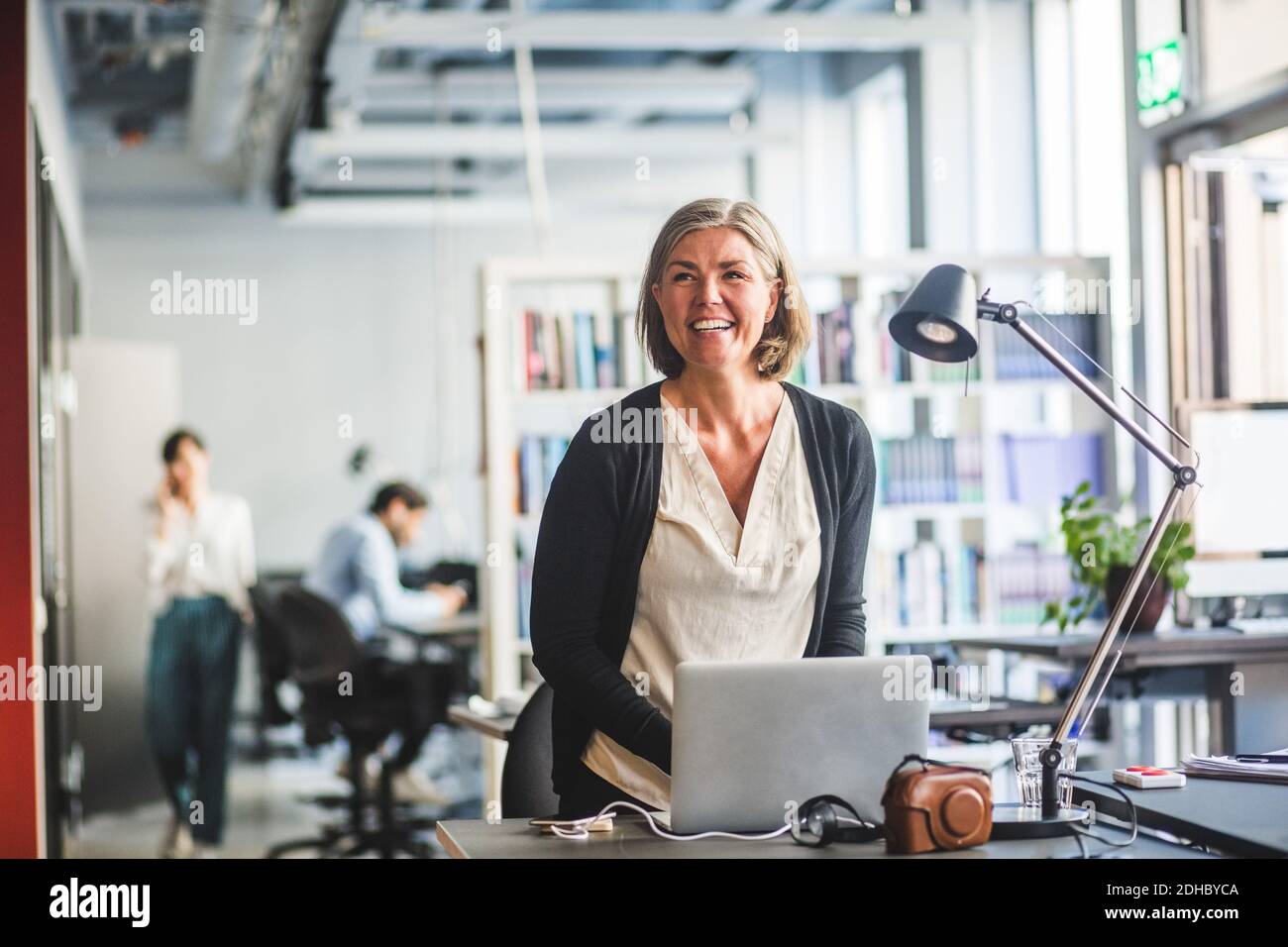 Donna d'affari felice e creativa che guarda via mentre si trova alla scrivania dentro ufficio Foto Stock