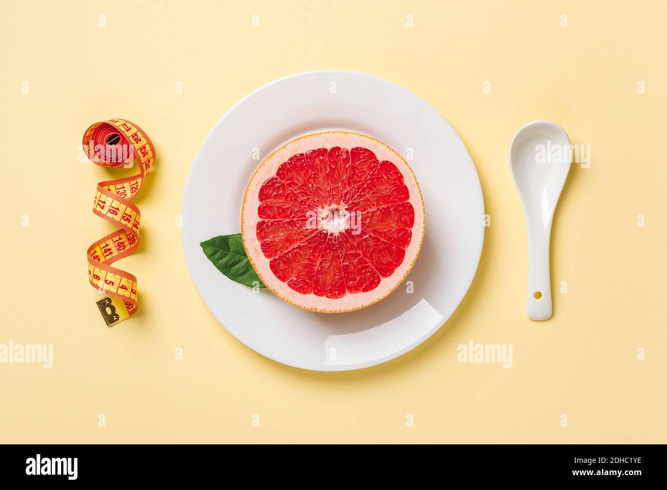 Metà di pompelmo succoso maturo su un piatto bianco, cucchiaio bianco e nastro di misurazione su sfondo giallo pastello. Dieta dimagrante, controllo del peso corporeo. Foto Stock
