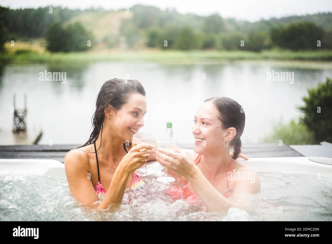 Sorridi le amiche che tostano gli occhiali da vino mentre ti godi nella vasca idromassaggio contro il lago Foto Stock