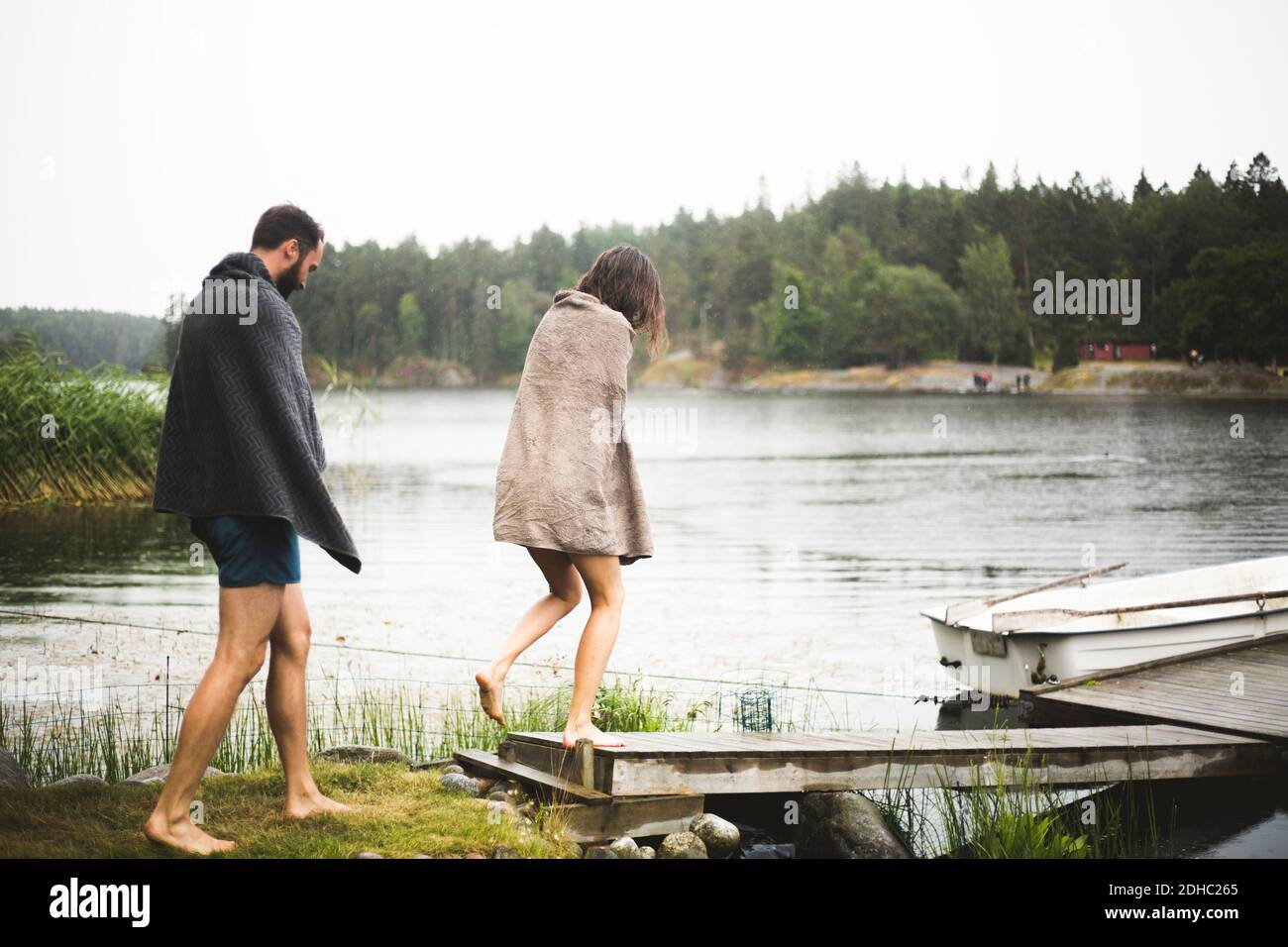 Amici maschili e femminili avvolti in asciugamani camminando verso il molo sul lago durante la fuga del fine settimana Foto Stock