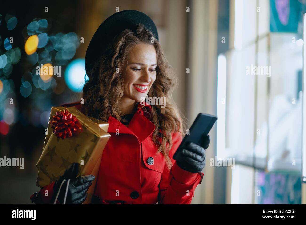 Divertimento invernale. Felice shopper donna di mezza età in cappotto rosso e berta nera con regali e smartphone che acquista online fuori in città in serata. Foto Stock