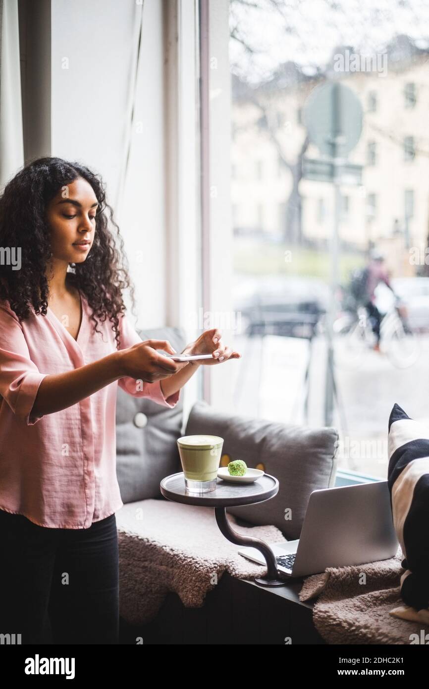 Giovane influencer femmina fotografando tè e snack sul tavolo attraverso smartphone in ufficio creativo Foto Stock
