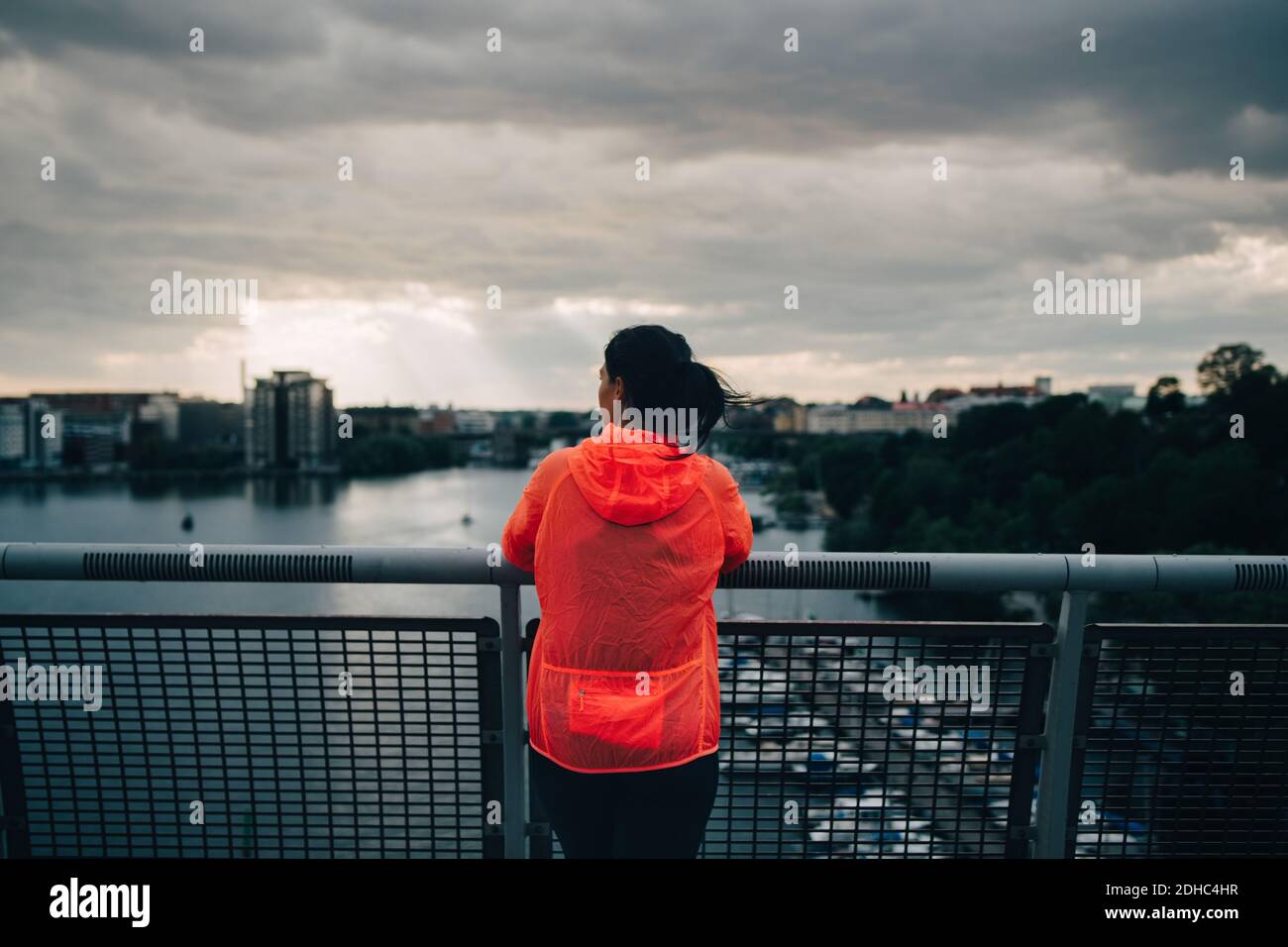 Vista posteriore dell'atleta femminile con impermeabile che guarda alla città mentre si è in piedi sul ponte pedonale sul mare durante il tramonto Foto Stock