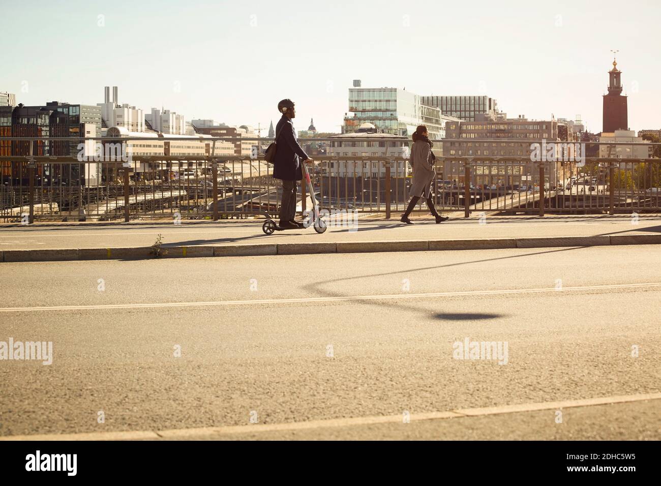 Vista laterale completa di un giovane uomo che corre in bicicletta elettrica da donna che cammina sul ponte in città Foto Stock