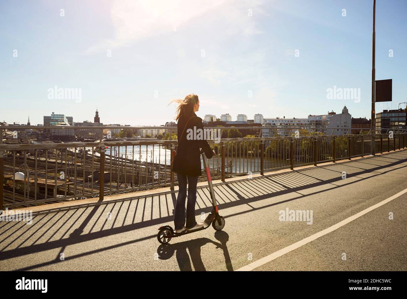 Per tutta la lunghezza del pendolari femmina che guidano lo scooter elettrico a spinta ponte in città contro il cielo Foto Stock