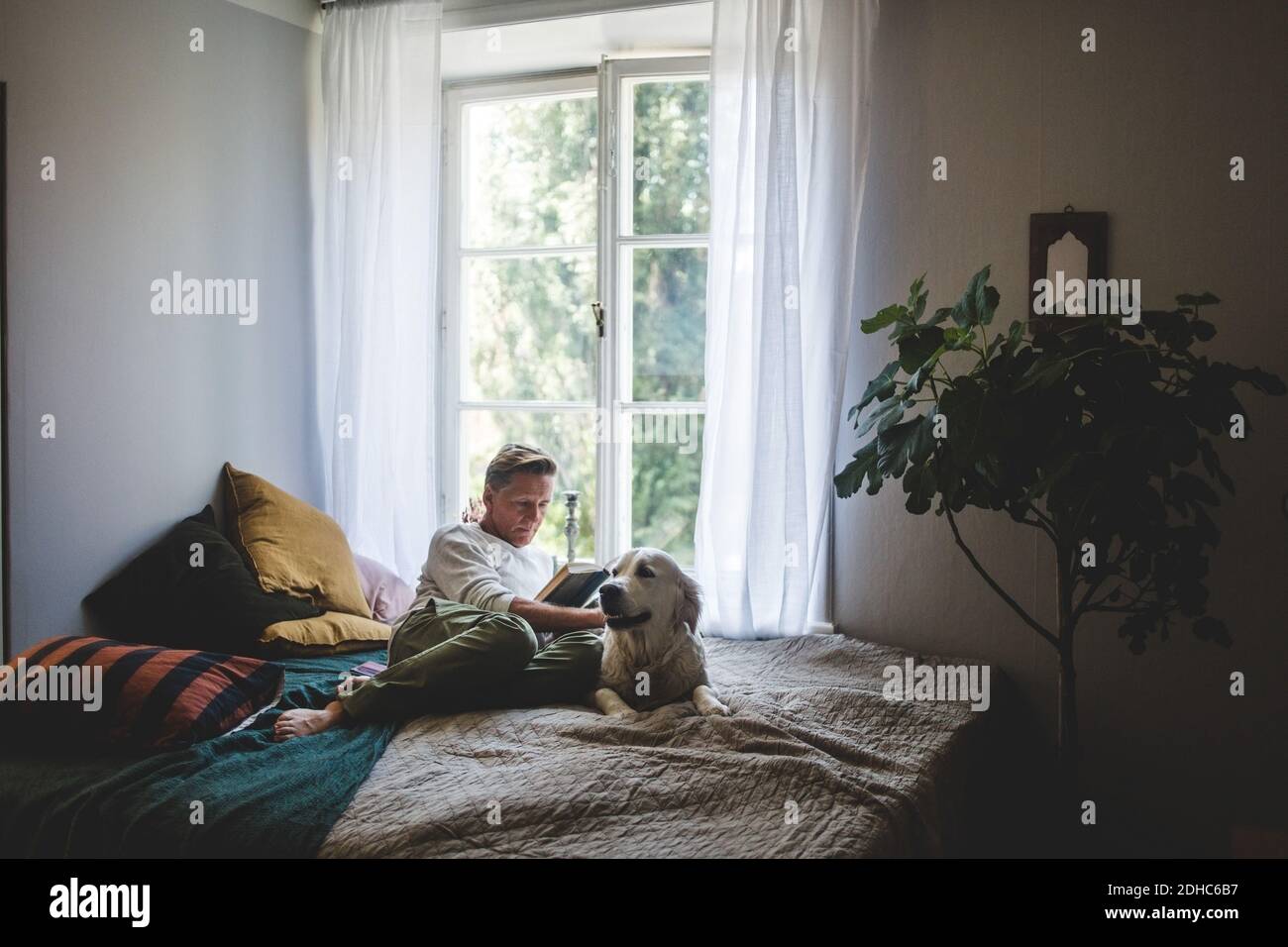 Lunghezza completa del libro di lettura dell'uomo in pensione mentre riposa con cane a letto a casa Foto Stock