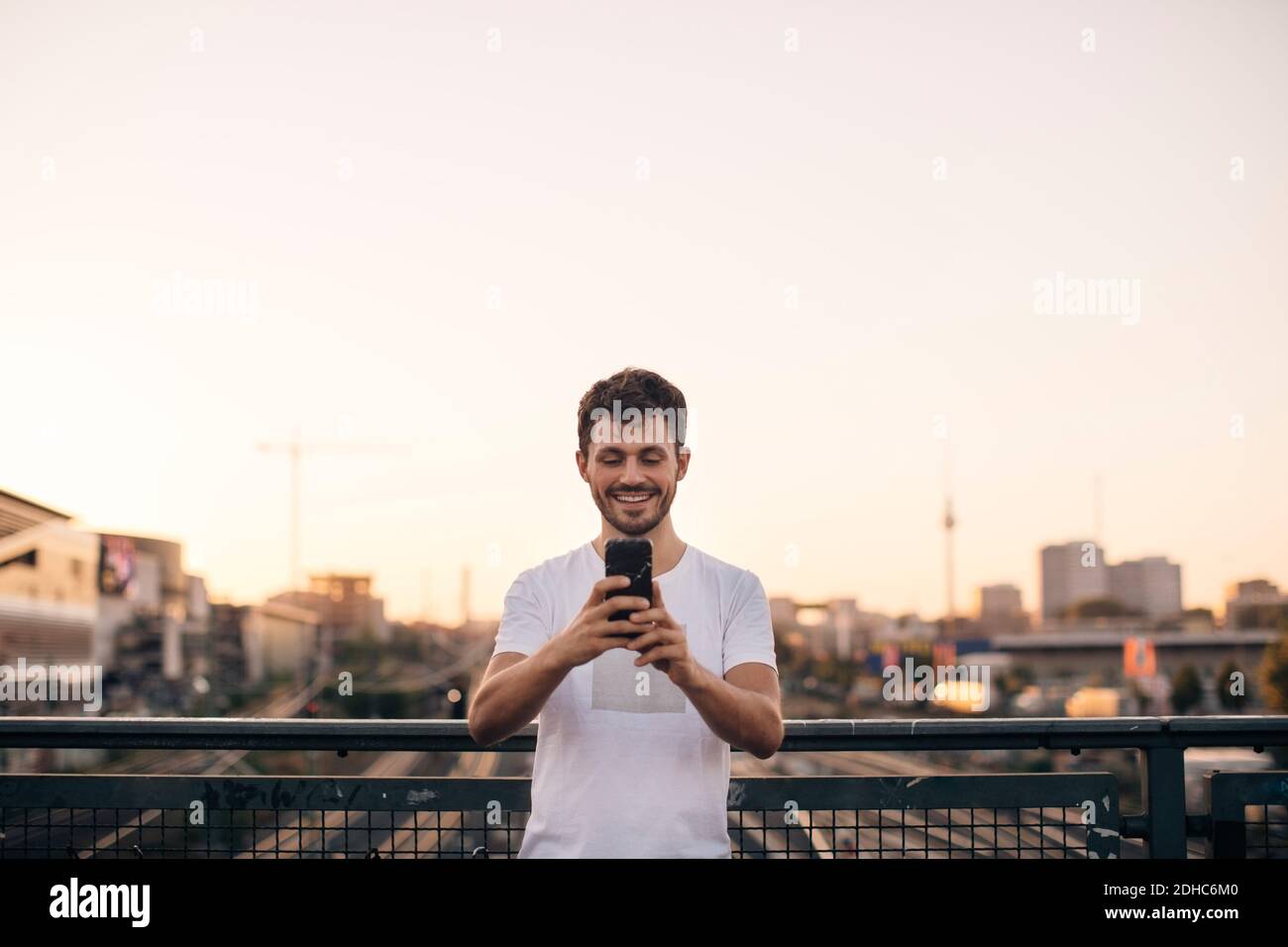 Sorridente giovane uomo che usa il telefono cellulare mentre si sta in piedi sul ponte contro il cielo limpido durante il tramonto Foto Stock