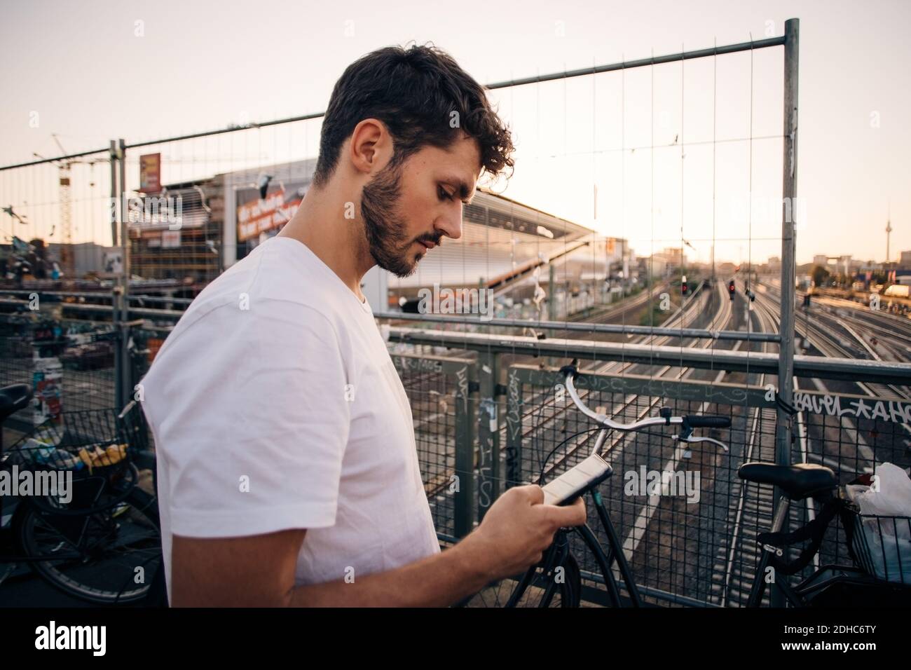 Vista laterale di un giovane uomo che utilizza il telefono cellulare mentre è in piedi sul ponte sopra i binari ferroviari in città Foto Stock