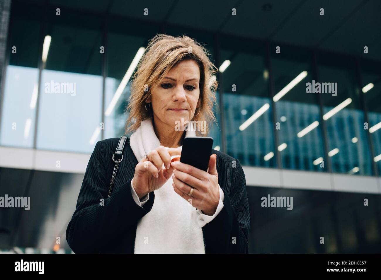 Vista ad angolo basso di una donna d'affari con capelli biondi con Smart telefono mentre si trova contro un edificio moderno in città Foto Stock