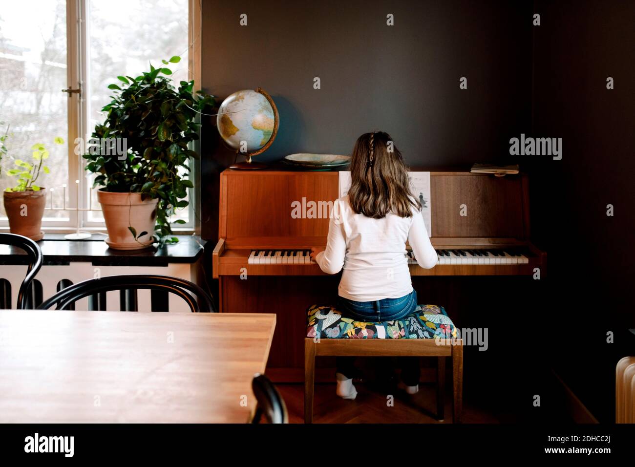 Vista posteriore della ragazza che suona il pianoforte mentre si siede in soggiorno camera a casa Foto Stock