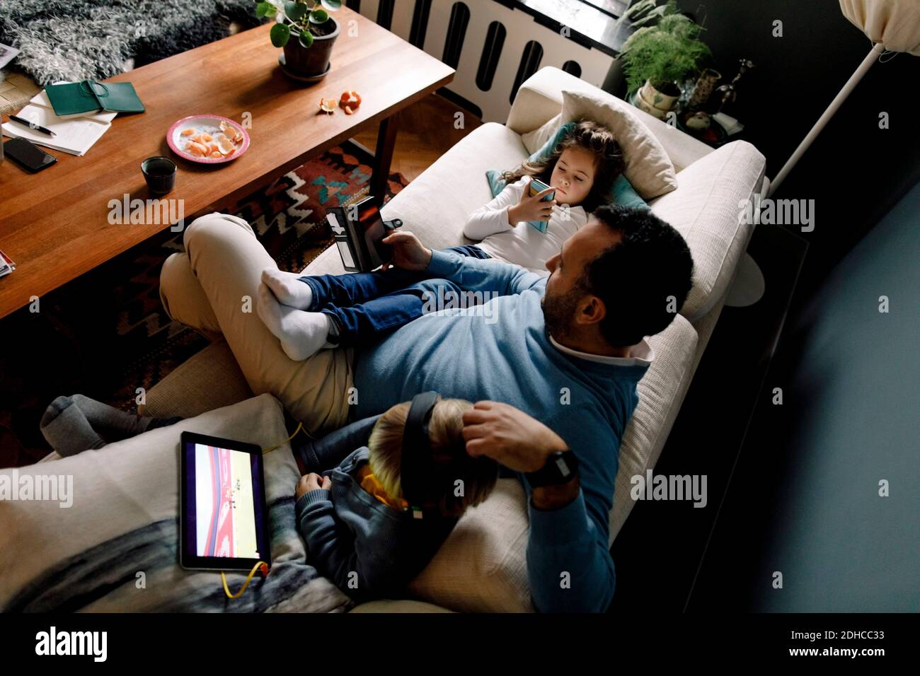 Vista ad alto angolo di padre e figlie utilizzando varie tecnologie sul divano in soggiorno a casa Foto Stock