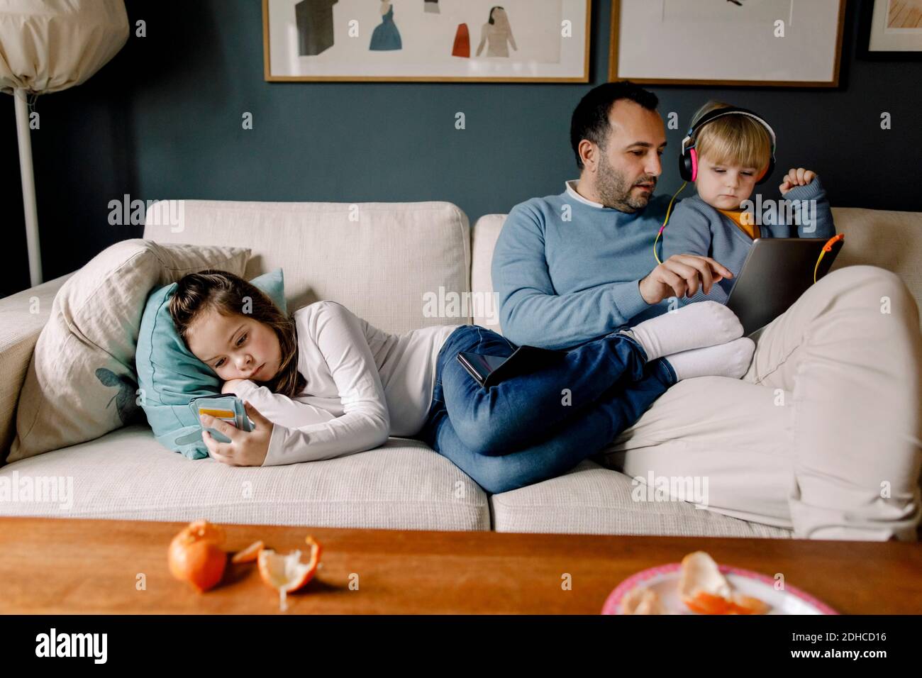 Ragazza che usa il telefono cellulare mentre padre mostra il tablet digitale a. sorella sul divano nel soggiorno di casa Foto Stock