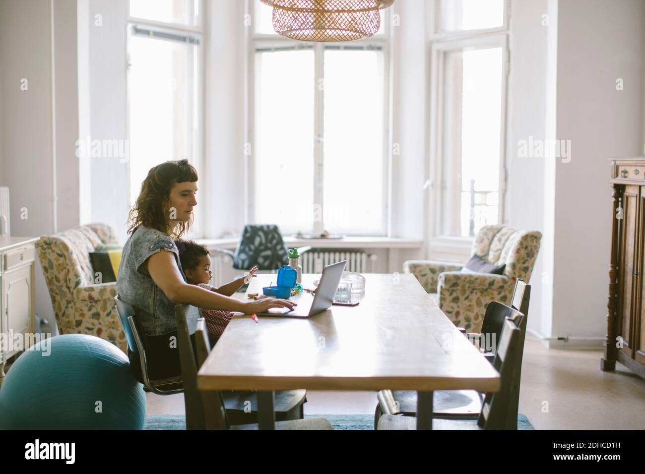Madre che usa il laptop mentre si siede con la bambina al tavolo in casa Foto Stock
