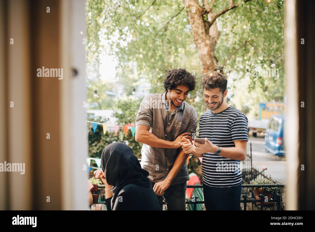 Felice giovane uomo che mostra il telefono cellulare ad un amico maschio mentre in piedi nel balcone durante la festa Foto Stock