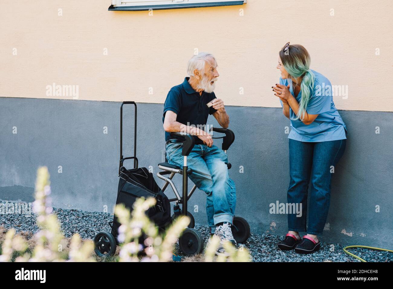 Sorridente infermiera che si aggrappò mentre si guarda l'uomo anziano seduto su sedia a rotelle contro il muro sul cortile posteriore Foto Stock