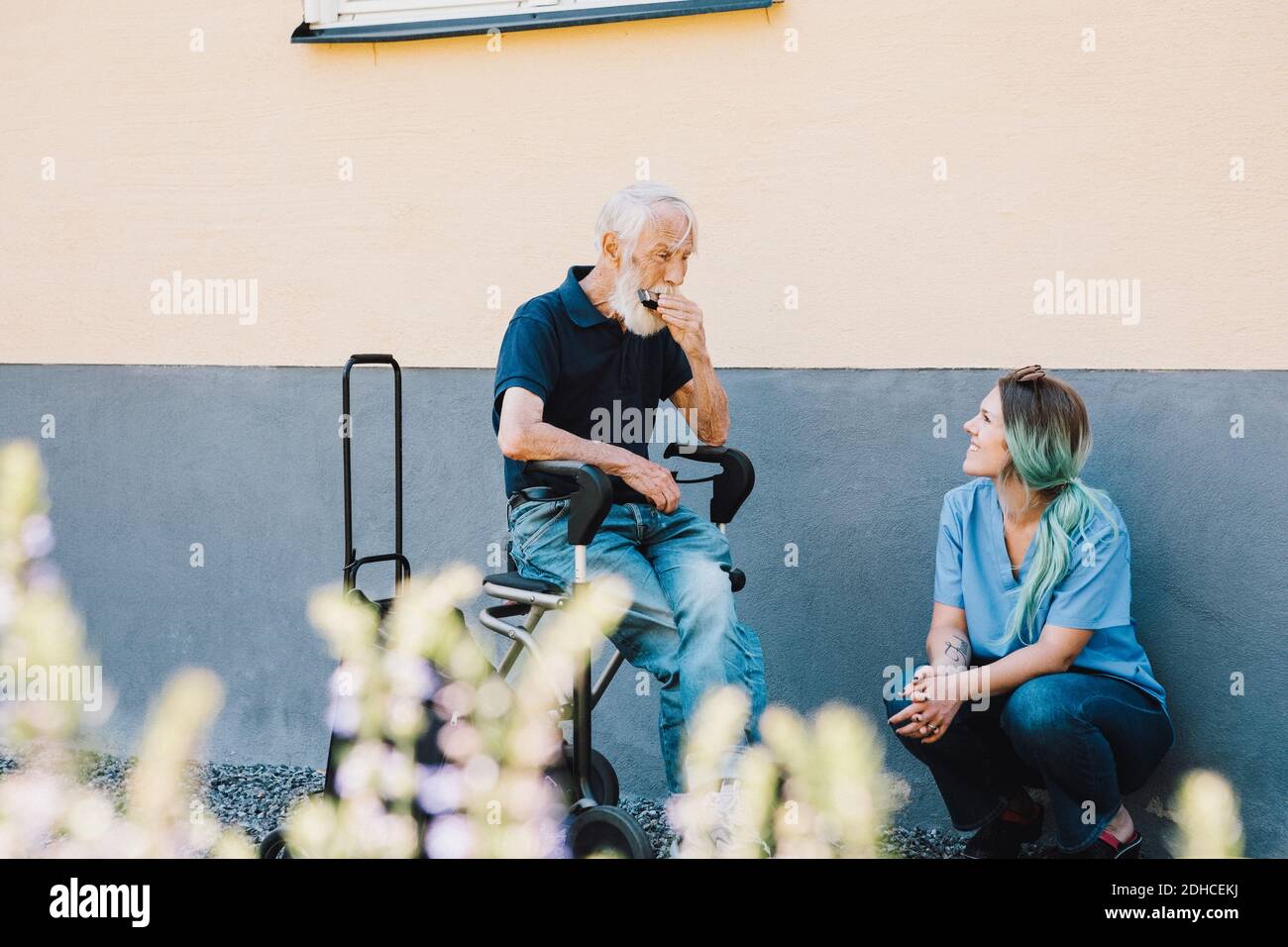 Femmina sorridente infermiera che guarda l'uomo anziano che gioca l'armonica contro muro sul cortile posteriore Foto Stock