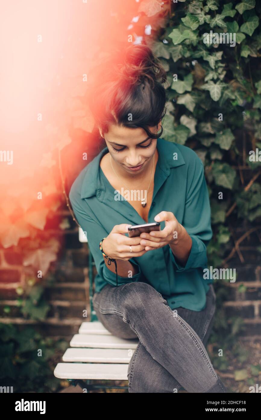 Giovane donna che usa il telefono cellulare mentre si siede sulla sedia cortile Foto Stock