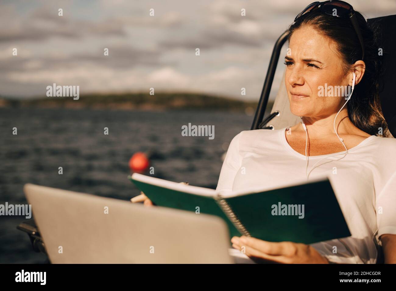 Donna premurosa con un notebook che tiene un diario mentre si guarda lontano contro lago Foto Stock