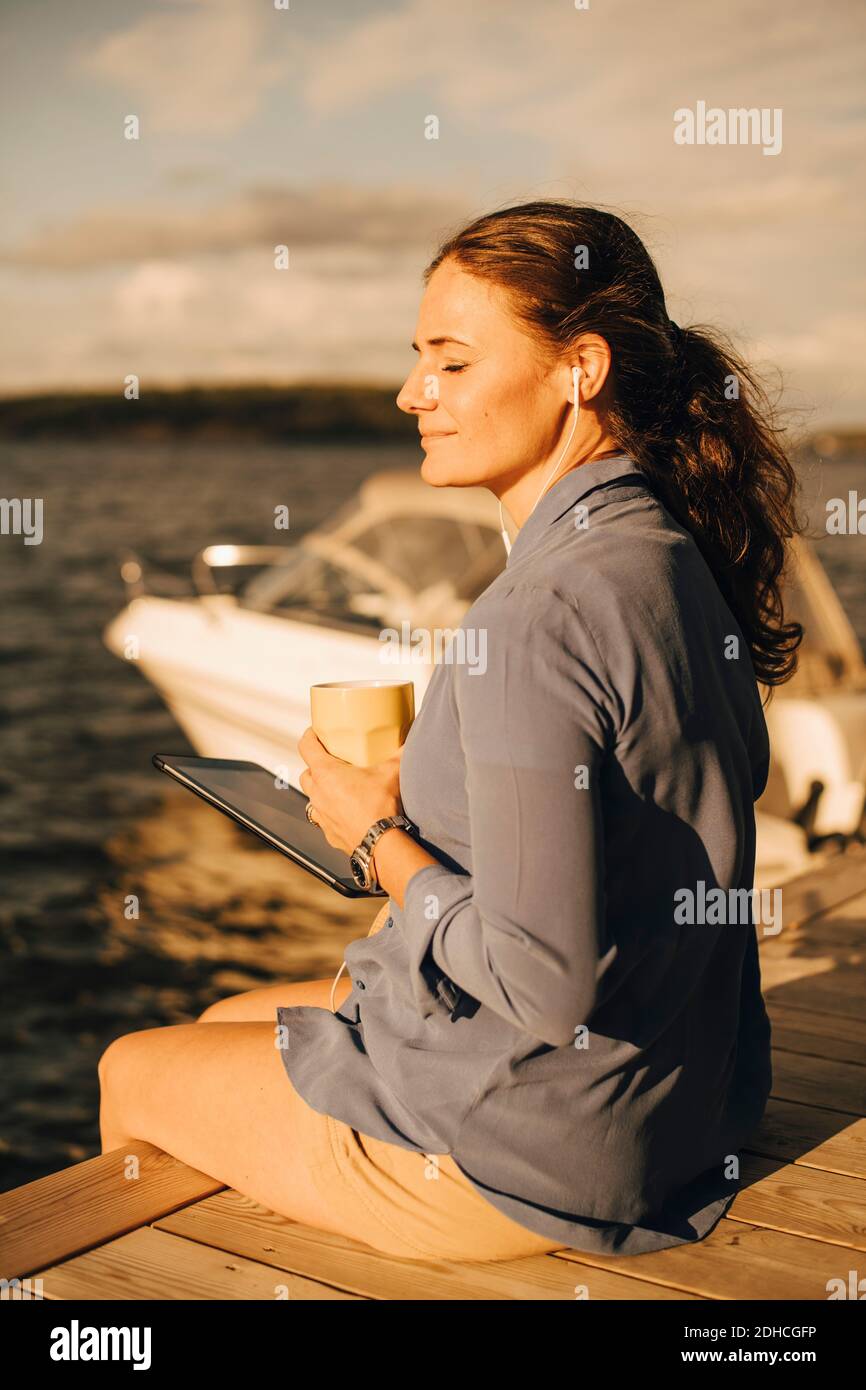 Donna matura sorridente che ha caffè mentre usa il tablet digitale AT patio contro il lago durante le vacanze Foto Stock