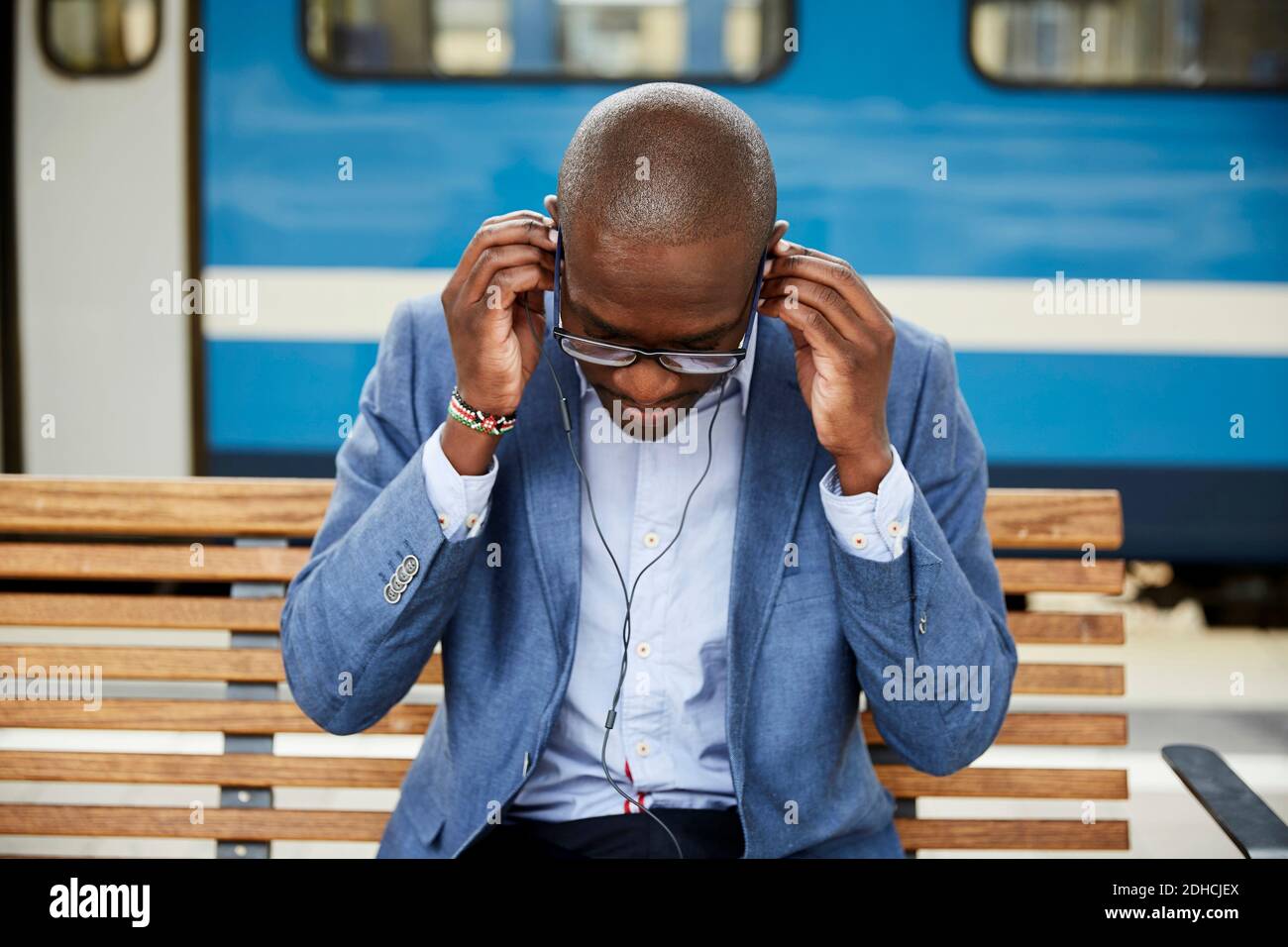 Uomo d'affari che regola le cuffie intrauricolari mentre si siede alla stazione ferroviaria Foto Stock