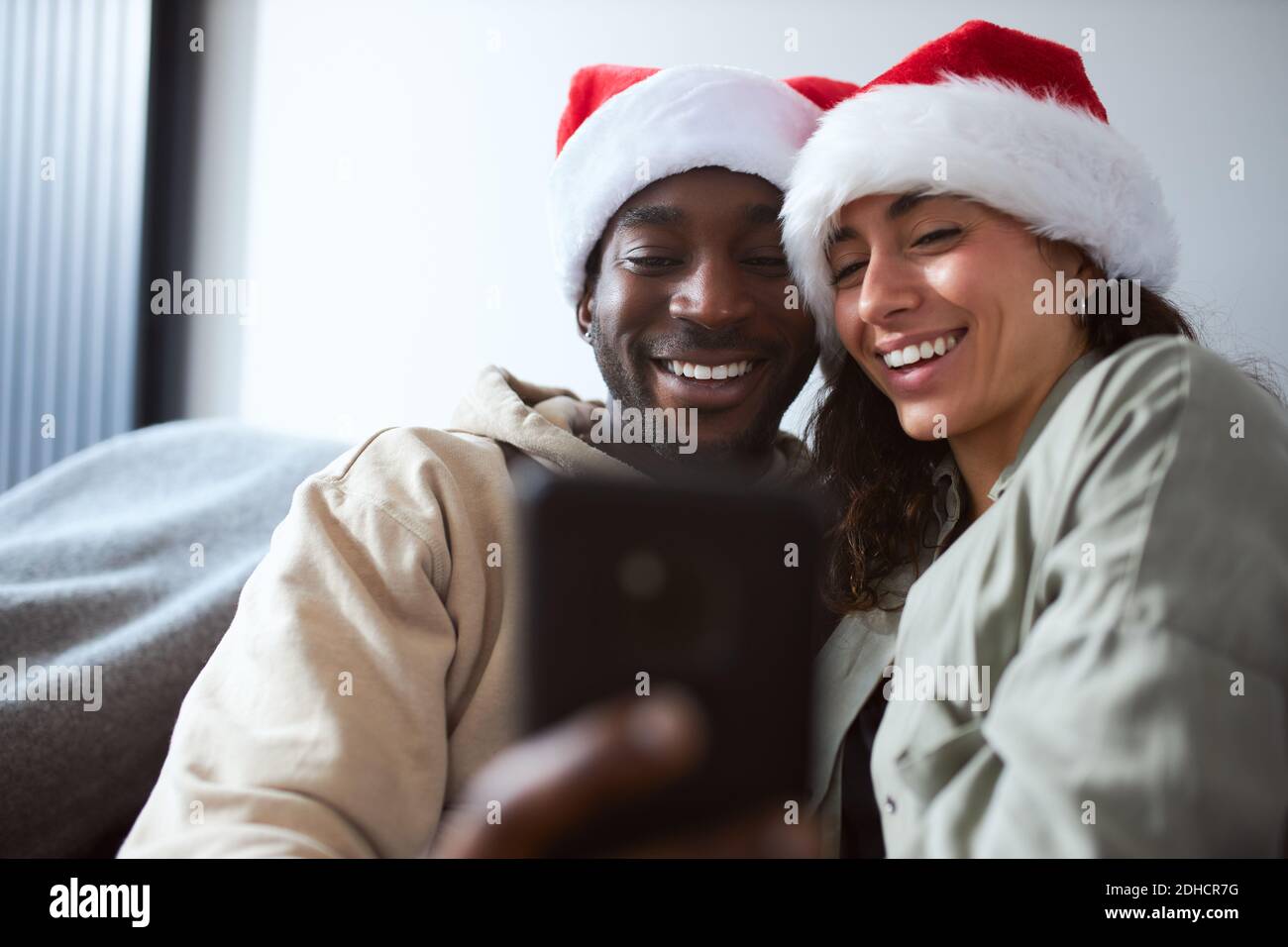 Giovane coppia di etnia mista che si rilassa sul divano a casa santa cappelli in posa per selfie natale sul telefono cellulare Foto Stock