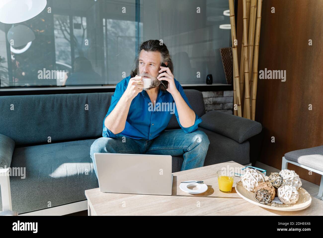 Uomo maturo con la barba che parla al telefono e beve un caffè seduto su un divano di fronte al portatile che guarda fuori dalla finestra. concetto di business Foto Stock