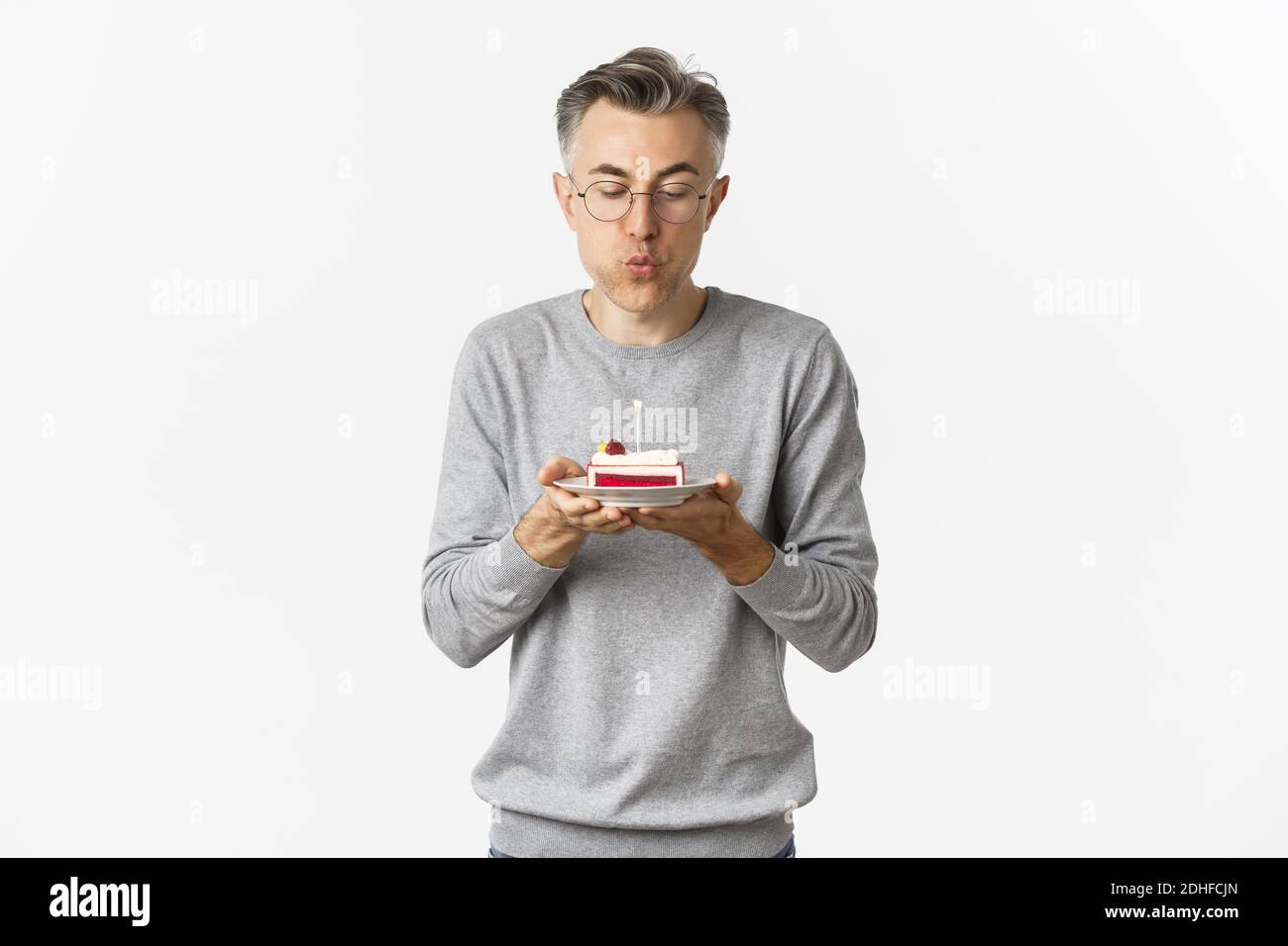 Ritratto di attraente uomo di mezza età in pullover grigio e bicchieri, celebrando il suo compleanno, soffiando una candela su torta b-day e. Foto Stock