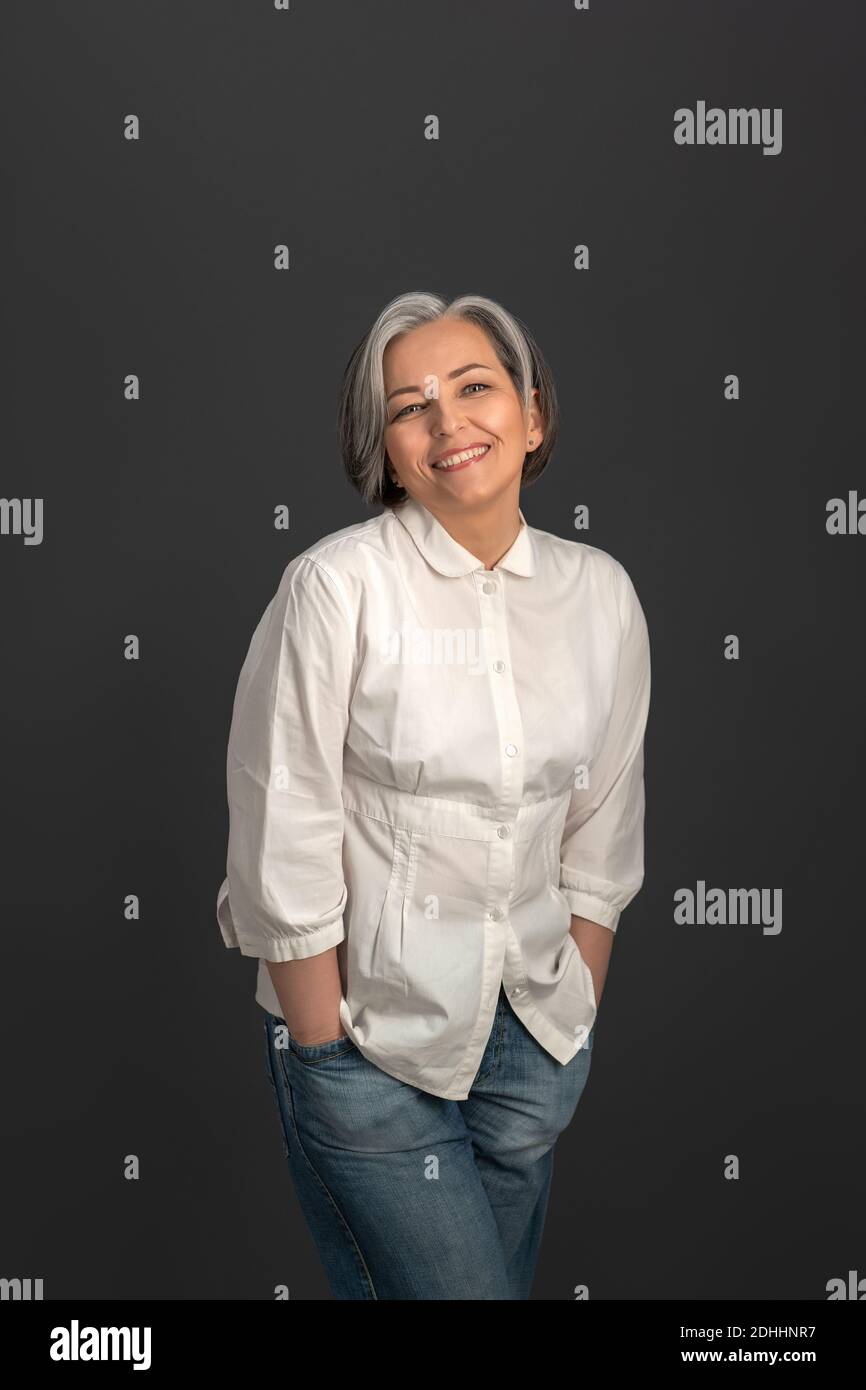 Senior grigio capelli affascinante donna aspetto felice indossare camicia bianca e jeans denim su fotocamera isolata su sfondo grigio. Ritratto di donna matura in Foto Stock