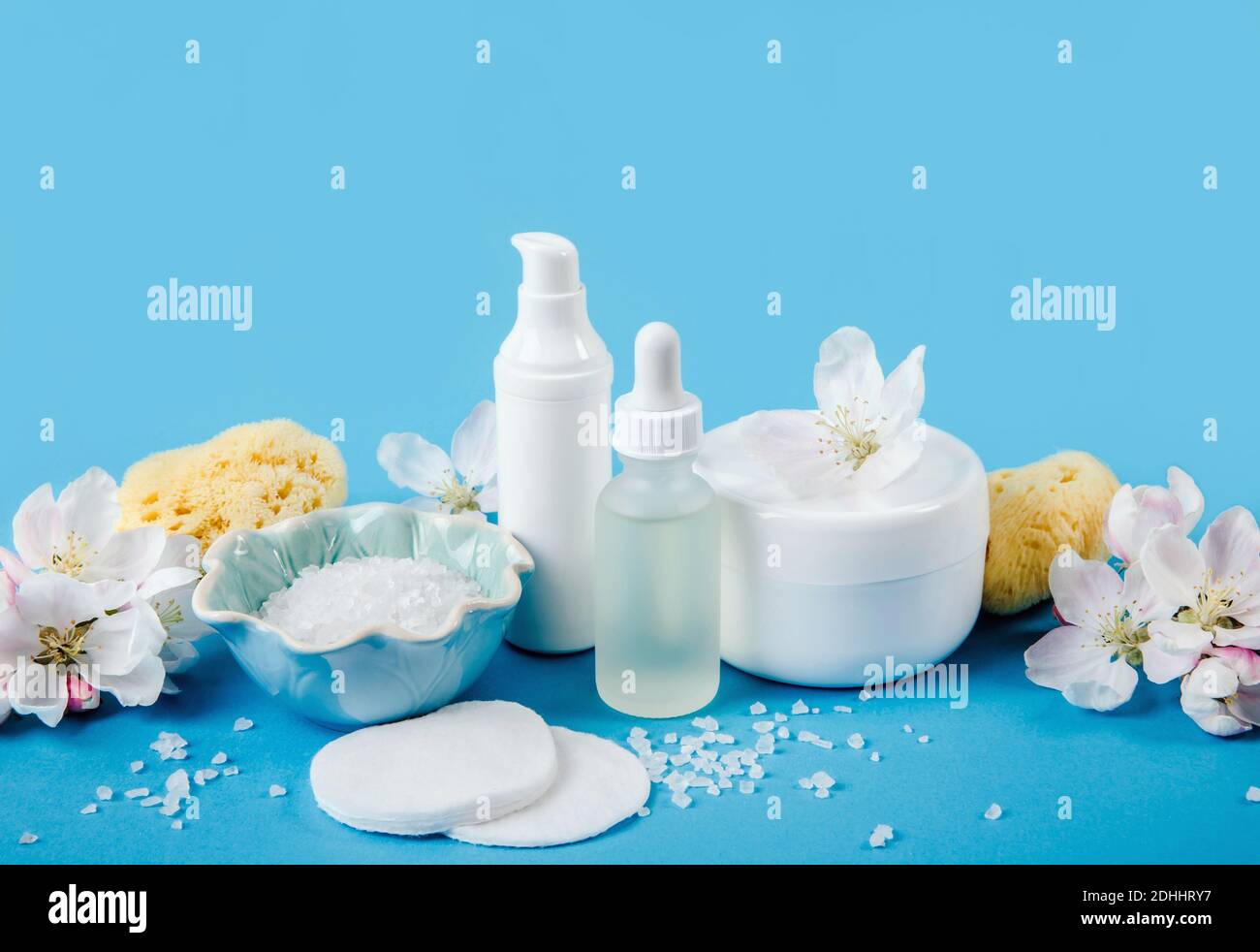 Vista laterale di contenitori di prodotti di crema di bellezza bianchi di varie dimensioni e forma con fiori di mela primaverile per la decorazione su sfondo azzurro. Foto Stock