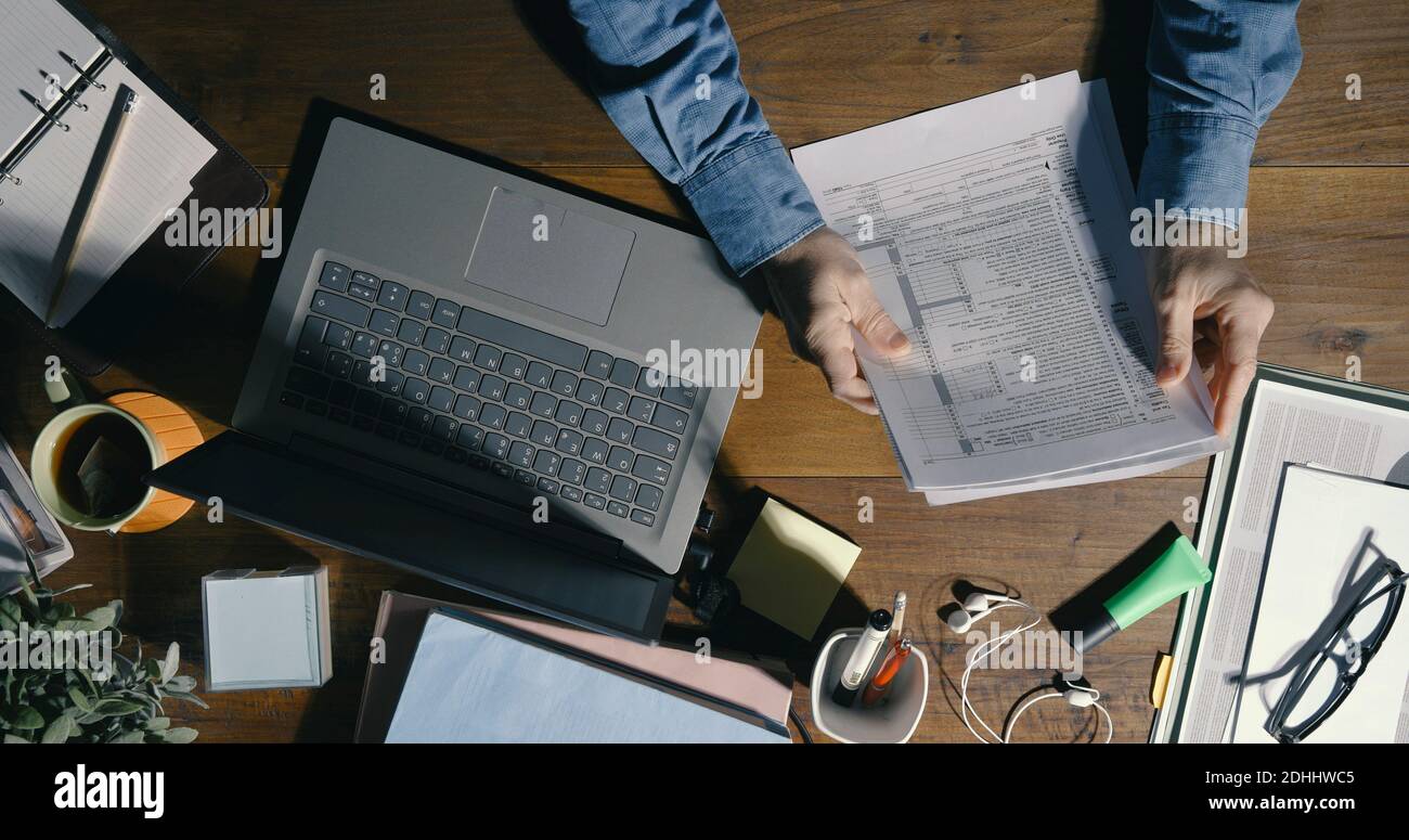 Uomo d'affari seduto alla scrivania dell'ufficio e che lavora, sta controllando i documenti e usando il suo laptop, vista dall'alto Foto Stock