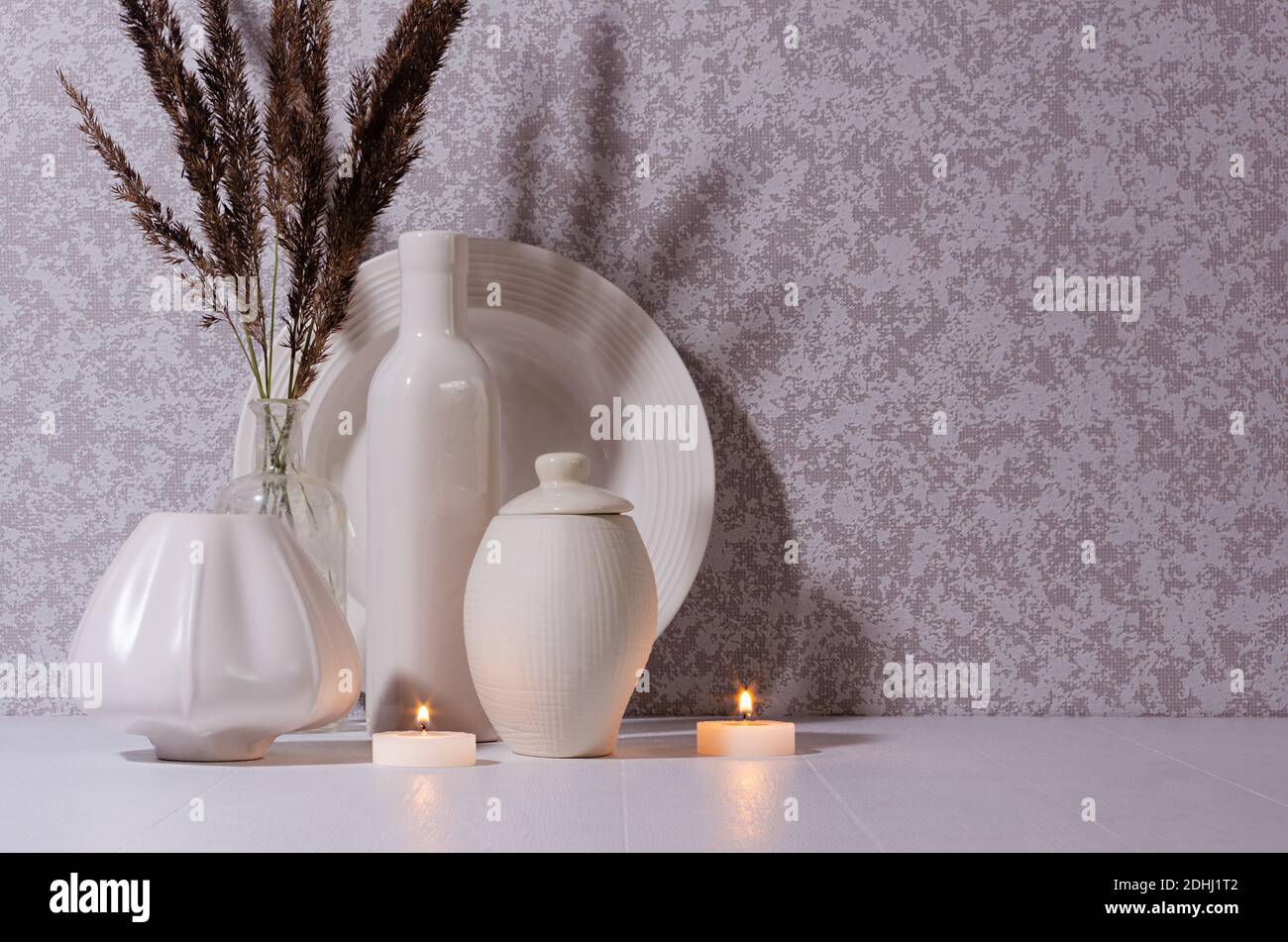 Elegante decorazione a casa di hygge in serata con candele brucianti, bottiglie di ceramica bianca, ciotola, piatto e bouquet di piante secche su tavola di legno bianco in moonligo Foto Stock