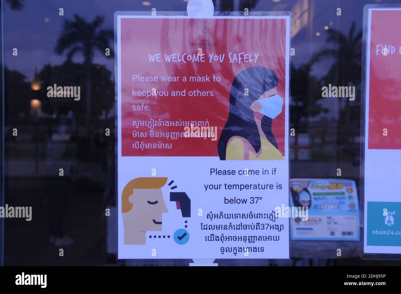 Un cartello di benvenuto bilingue COVID - 19, con riflesso tropicale, all'ingresso di una panetteria francese sul fiume durante la pandemia del coronavirus. Phnom Penh, Cambogia. © Kraig Lieb Foto Stock