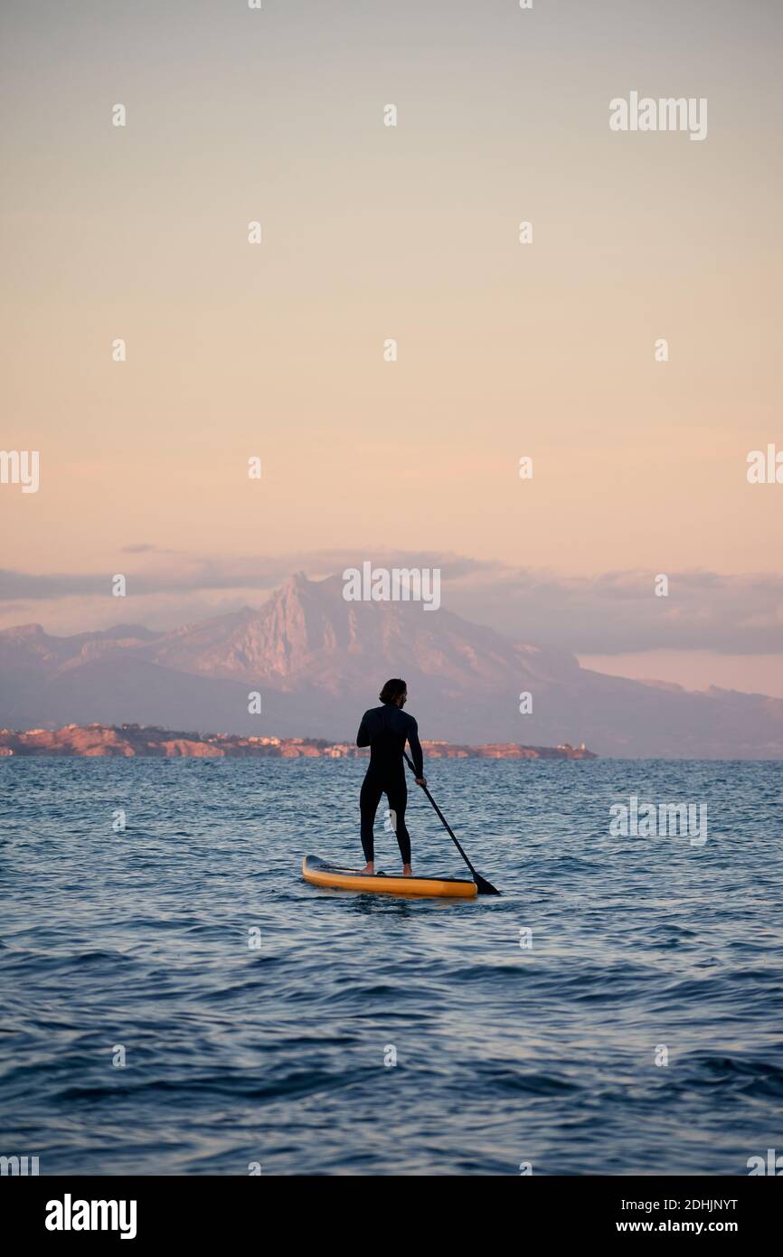 Surfista maschile in muta canottiera su tavola a pale in mare acqua sullo sfondo delle montagne al tramonto Foto Stock