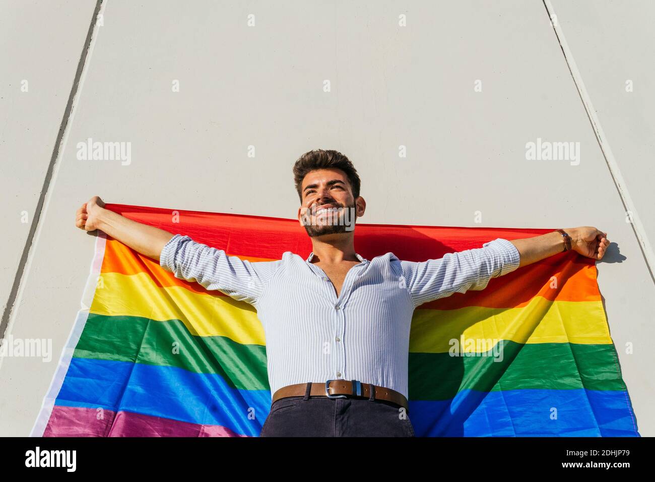 Da sotto allegro omosessuale maschile con arcobaleno LGBT bandiera in piedi su un muro di cemento sulla strada della città in giornata di sole Foto Stock