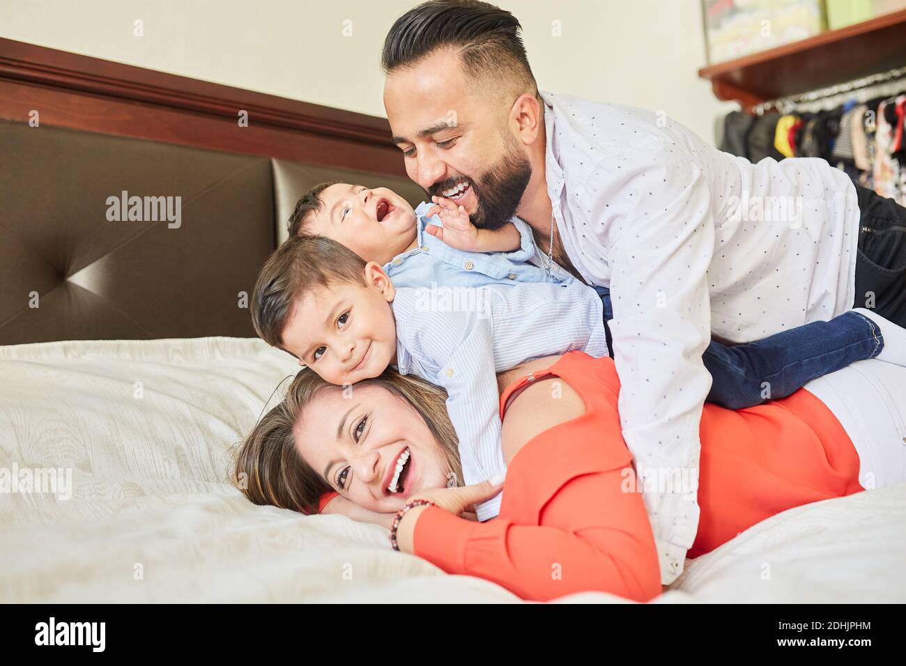 Una coppia ispanica positiva con dei piccoli figli carini che si riuniscono a letto e guardando la fotocamera mentre ti godi il passatempo a casa Foto Stock