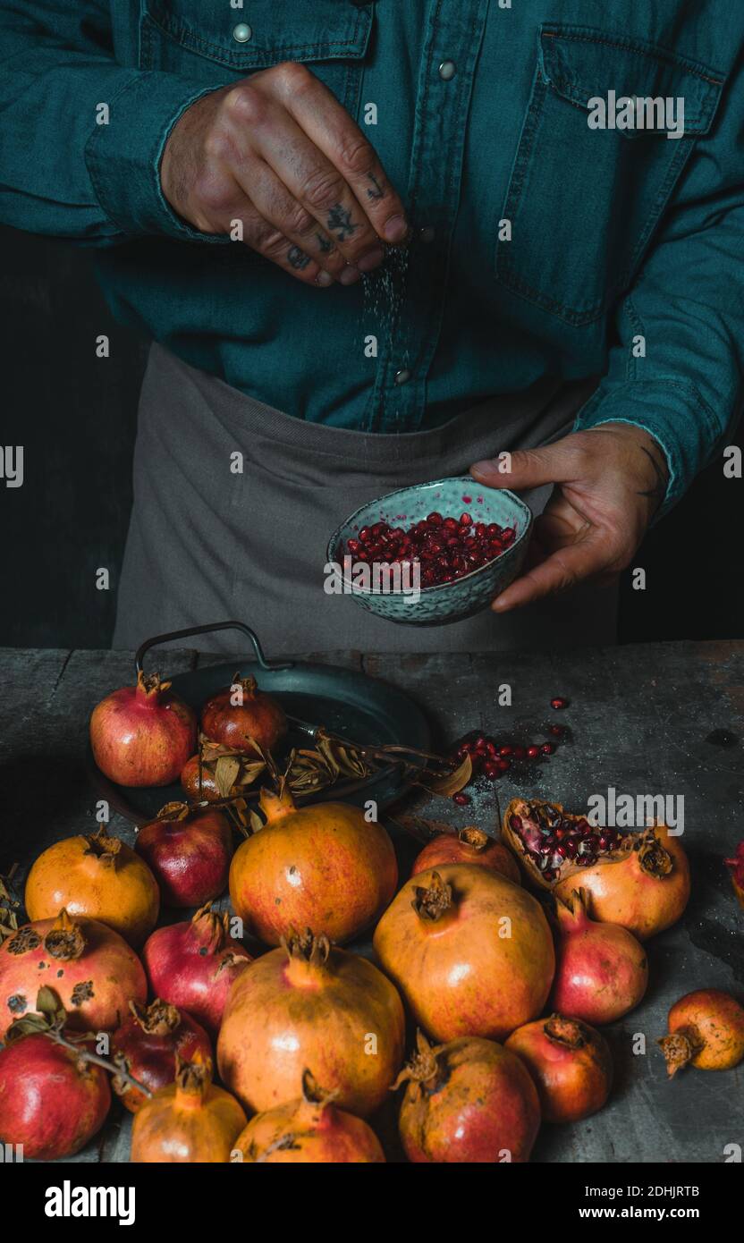 Angolo alto di cottura maschile che spruzzano lo zucchero nel recipiente con deliziosi semi di melograno maturi mentre si trova a tavola rustica Foto Stock