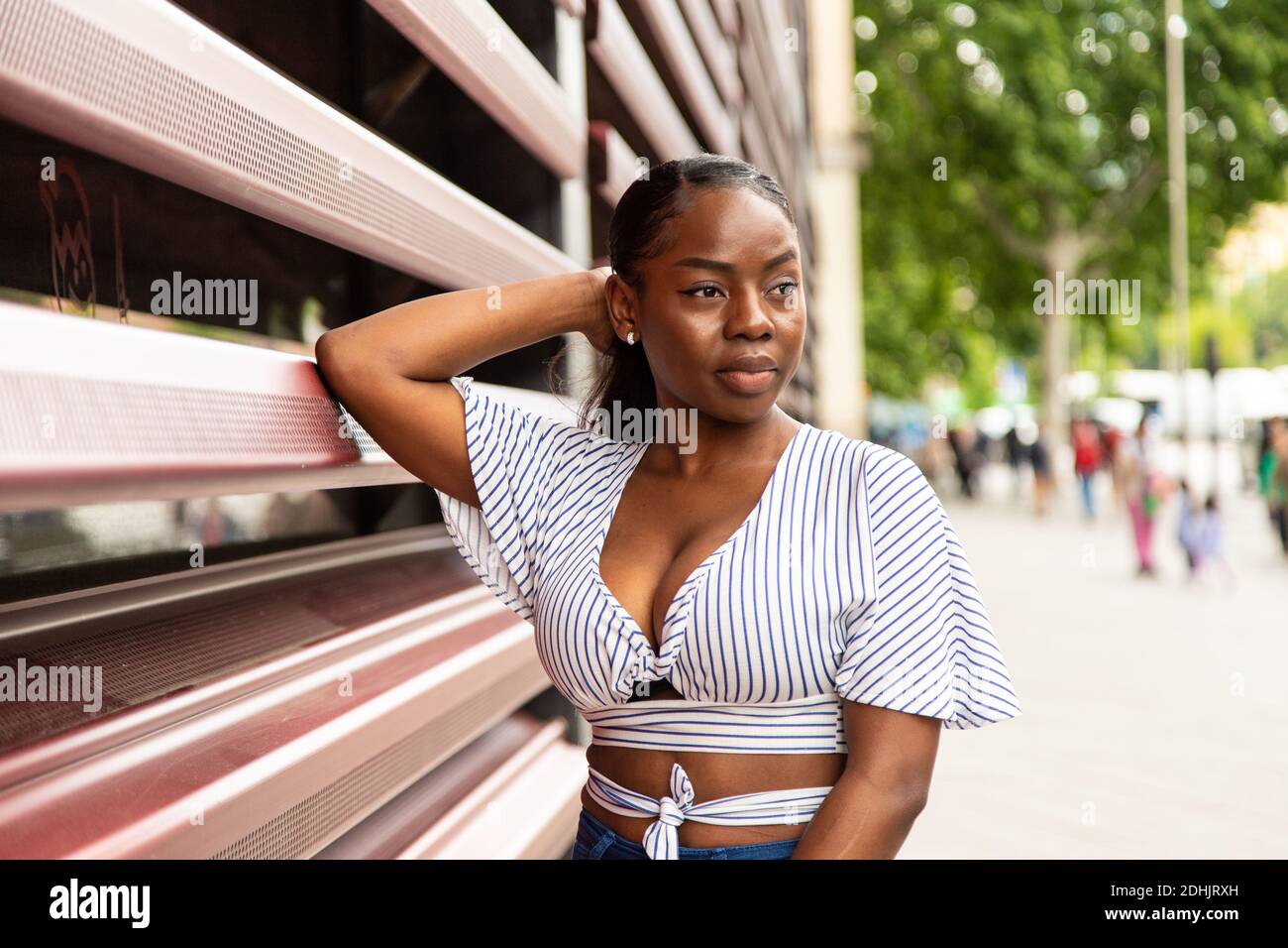 Moderna giovane donna afroamericana in elegante blusa a righe lontano mentre si trova vicino alla ferrovia di un edificio moderno sulla città street in estate da Foto Stock