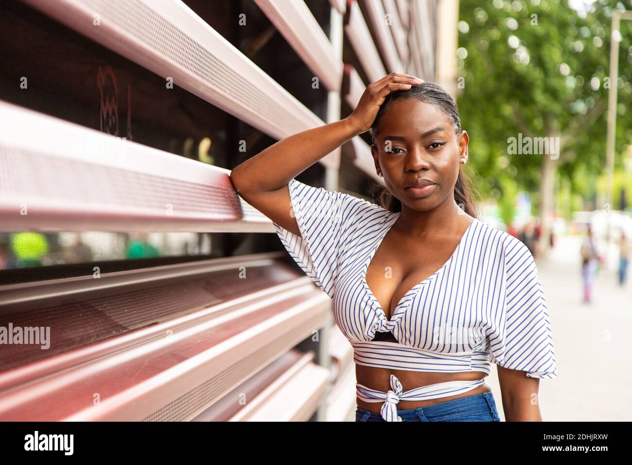Moderna giovane donna afroamericana in elegante blusa a righe a macchina fotografica mentre si è in piedi vicino alla ringhiera di edificio moderno su via della città in summ Foto Stock