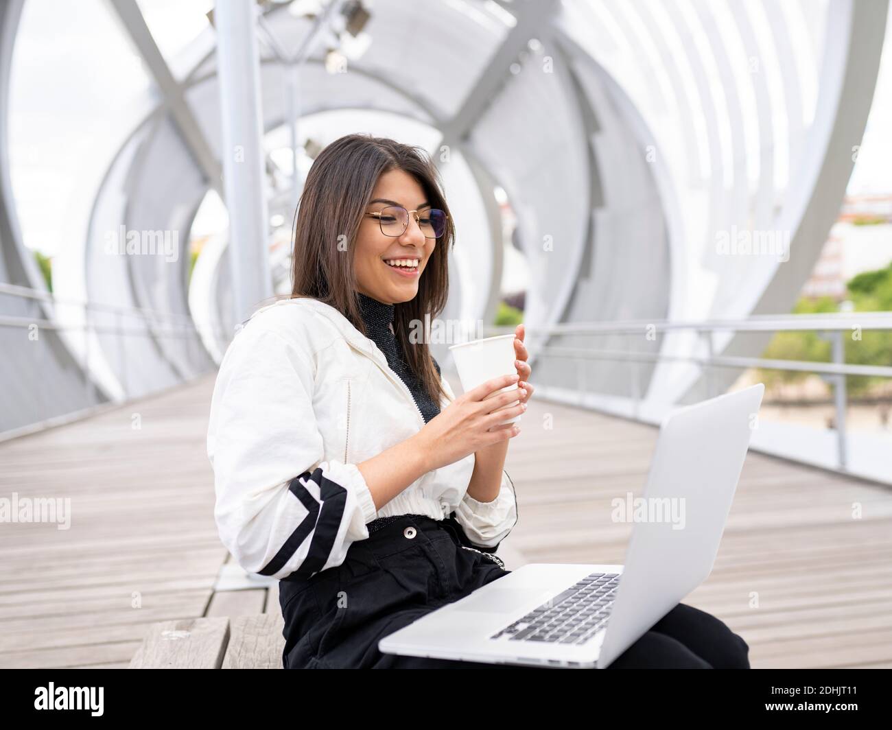Vista laterale di positivo moderno millenario etnico blogger femmina bere caffè da asporto e utilizzo di un computer portatile mentre si siede su un ponte chiuso oltre il fiume in Foto Stock