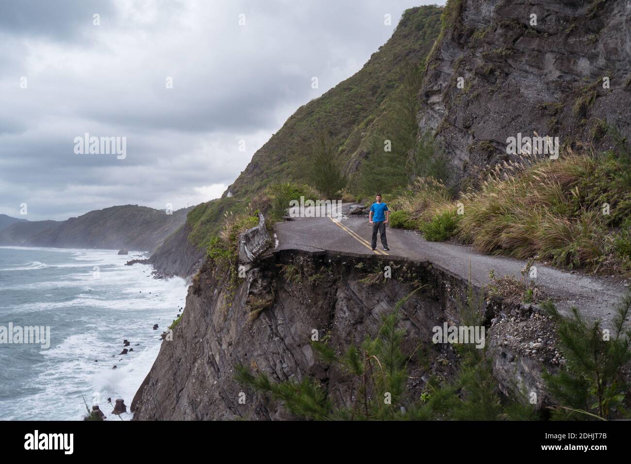 Irriconoscibile viaggiatore maschile distante in piedi sul bordo di asfalto distrutto strada che corre lungo la costa rocciosa ruvida vicino mare ondeggiante in tempo eccessivo Foto Stock