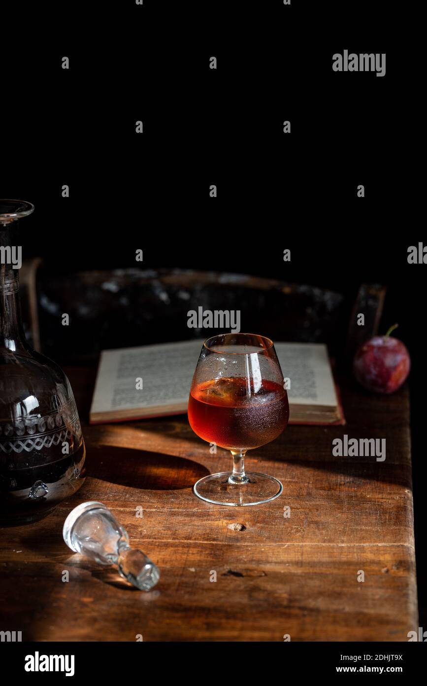 Bicchiere di cognac con cubetti di ghiaccio posti su tavolo di legno con decanter e prugna fresca Foto Stock