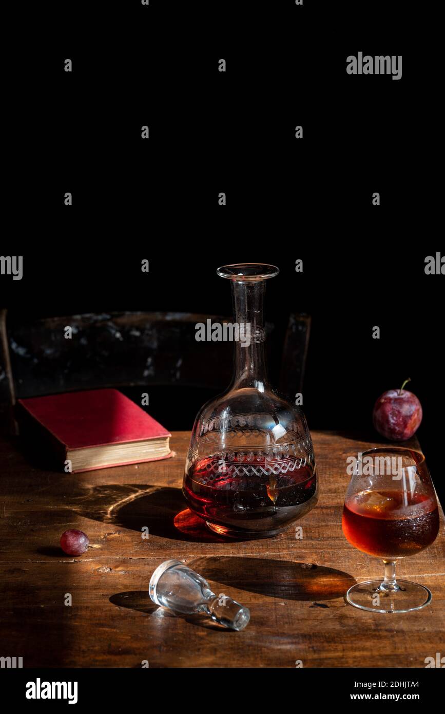Bicchiere di cognac con cubetti di ghiaccio posti su tavolo di legno con decanter e prugna fresca Foto Stock