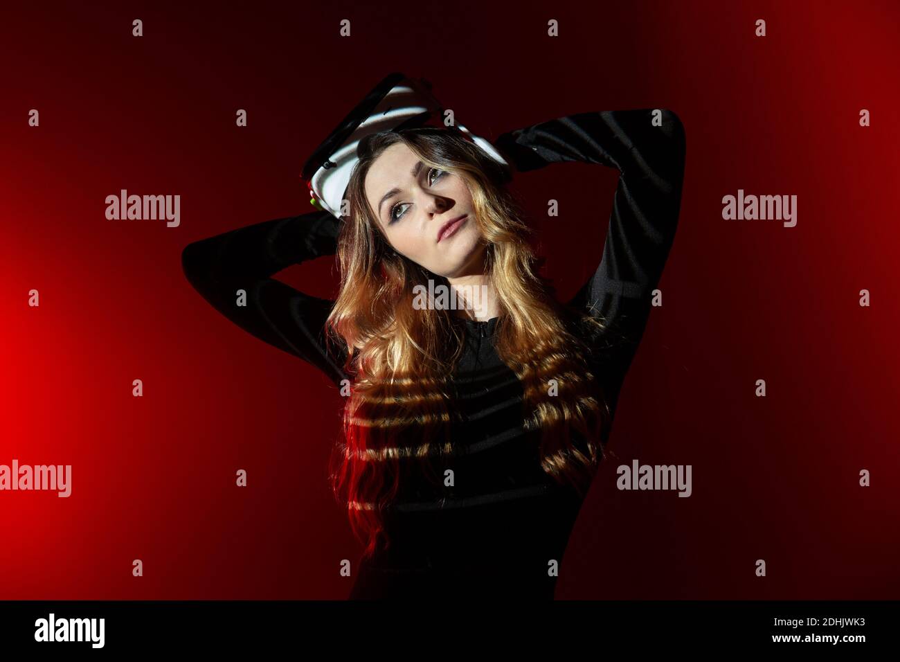 Giovane donna bionda che indossa una cuffia per realtà virtuale in piedi su uno sfondo colorato rosso da studio guardando lontano Foto Stock